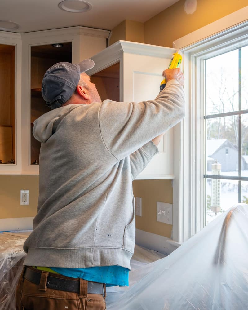 Man in a cap and gray hoodie running a bead of caulk along a white kitchen wall cabinet beside a window, counters covered in plastic