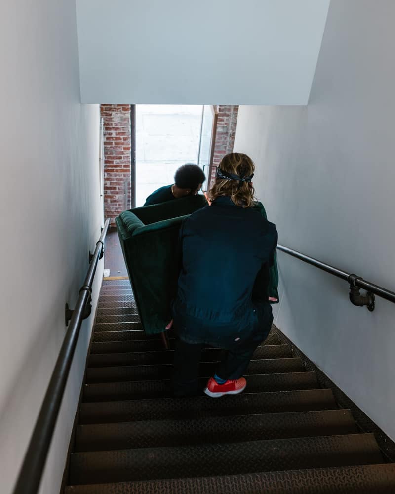 Two men carrying a green velvet sofa down a narrow stairwell between two handrails, toward a bright doorway set in brick