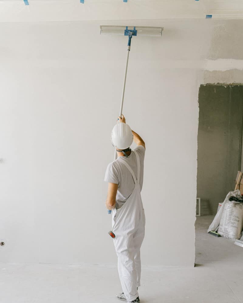 Painter in white overalls and a hard hat rolling a ceiling with an extension pole in an empty room