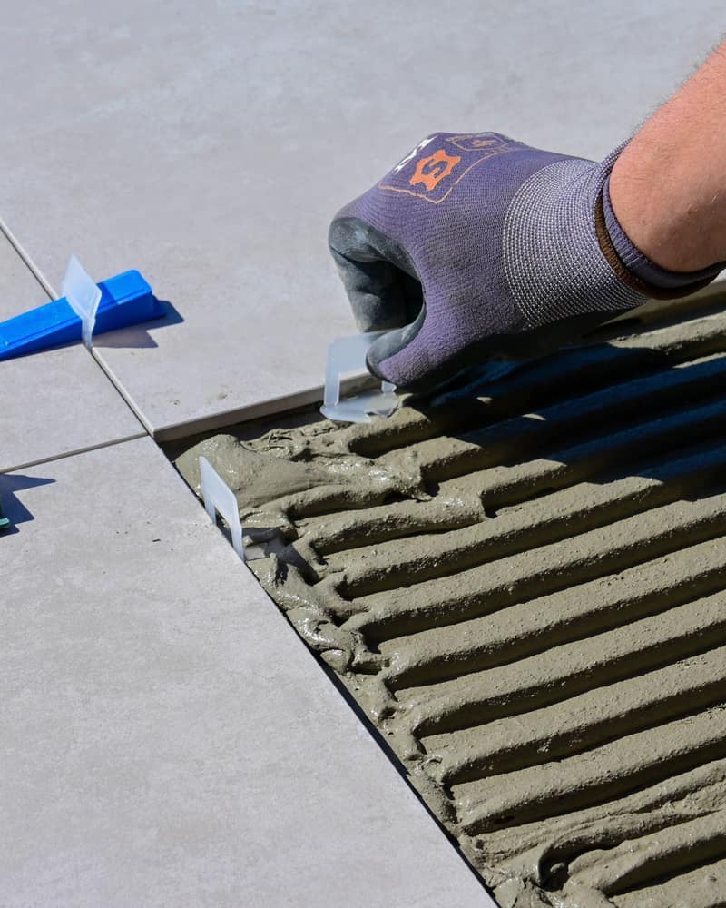 Gloved hand setting a leveling clip between large gray tiles pressed into combed thinset, with a blue leveling wedge beside it