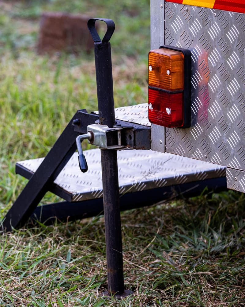 Drop-leg jack stand clamped to the corner of a diamond-plate trailer beside its amber and red tail light, over a fold-down step in the grass