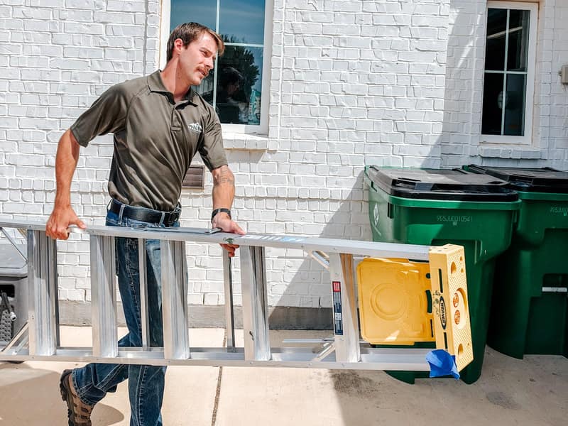 Jaden Christian, owner of Trailhead Handyman, carrying an aluminum stepladder past a white brick house