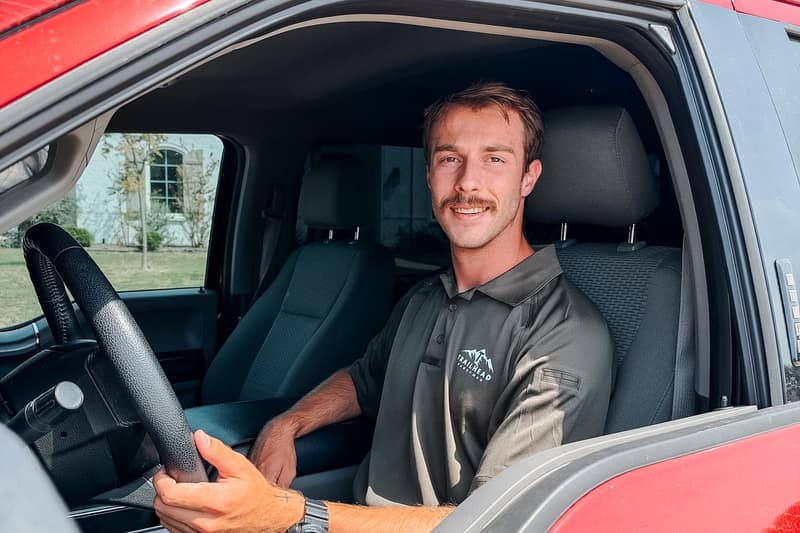 Jaden Christian, owner of Trailhead Handyman, in a gray Trailhead polo, smiling from the driver seat of a red pickup truck