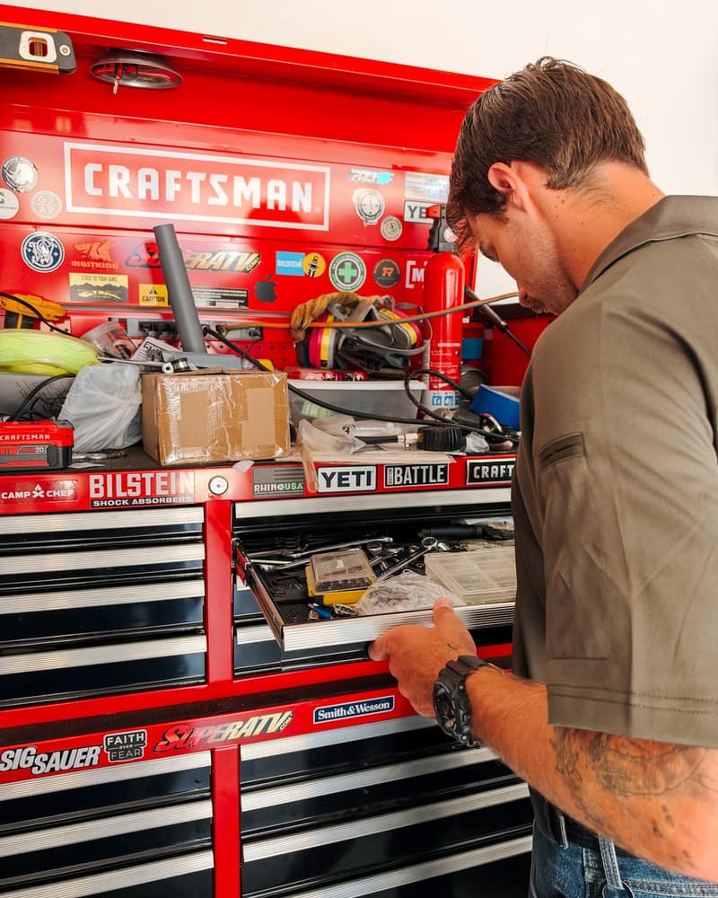 Jaden Christian, owner of Trailhead Handyman, working on a part at a red rolling tool chest in a garage shop