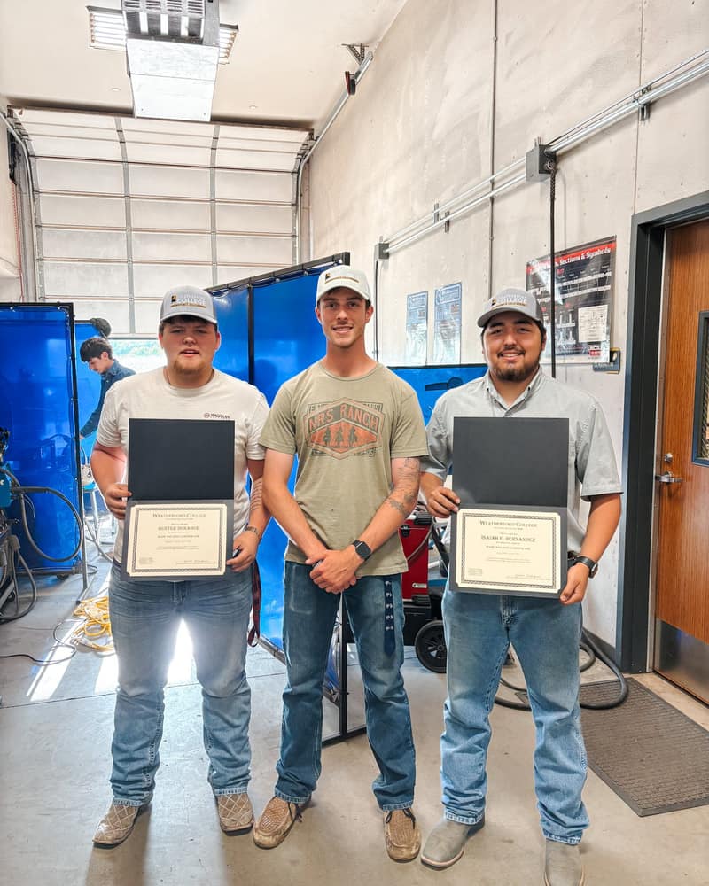 Jaden Christian, owner of Trailhead Handyman and a welding instructor, standing between two students holding certificates in the Weatherford College welding shop